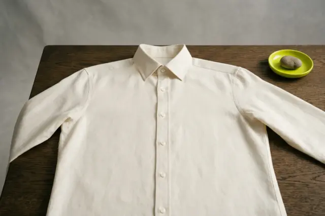 An impeccably ironed cream linen button-down shirt, fully buttoned and open flat, displaying its precise collar, smooth placket, and perfectly aligned shoulder seams on a dark oak wooden table.