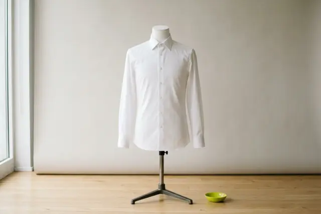 A perfectly tailored white cotton dress shirt smoothed tautly onto a headless white dress form, highlighting the precise shoulder seams and a neat, uncreased chest on a light wooden floor.
