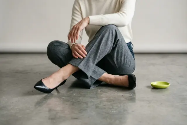 A person's legs from mid-thigh, wearing gray wool trousers and black leather pumps, elegantly crossed while seated on a polished concrete floor.