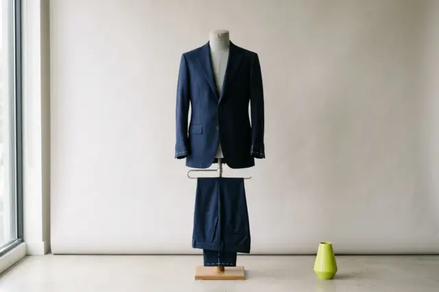 A navy wool suit jacket and matching trousers are precisely draped on a light gray dress form, with white tailor's chalk marks subtly indicating alteration points on the sleeve and pant hem.