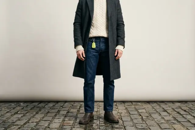 A person from shoulders down, wearing a charcoal wool overcoat over a cream chunky knit sweater, with sleeves covering wrists and arms slightly bent, standing on a cobblestone street.