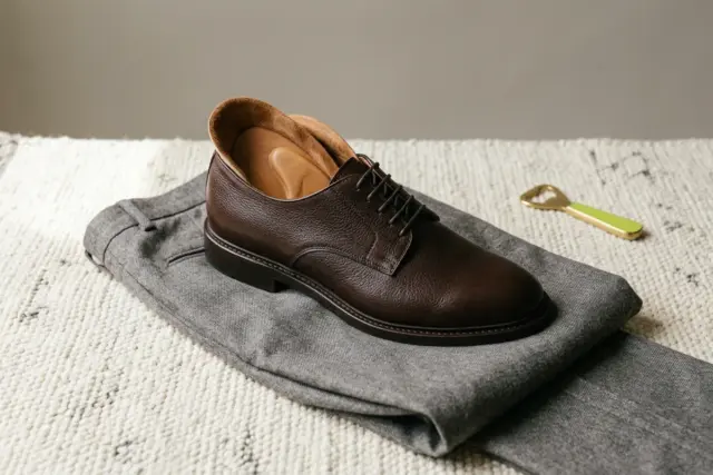 A single dark brown leather derby, angled to reveal its plush interior lining and a supportive insole, rests on a softly folded gray flannel trouser leg atop a textured light-colored rug.