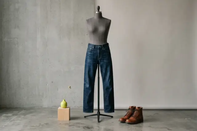 Dark-wash straight-fit denim jeans hang on a headless dress form, the waist and seat gently contoured, with cognac leather boots placed on the floor nearby, against a grey concrete wall.