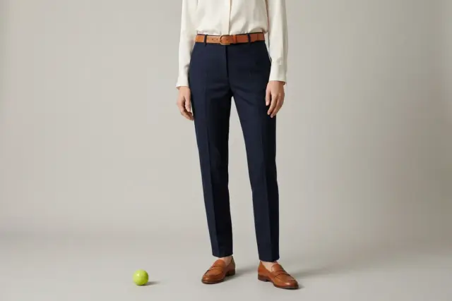 A person, photographed from shoulders down against a warm beige studio backdrop, wears perfectly tailored navy wool trousers, an ivory silk blouse, a cognac leather belt cinching the waist, and matchi