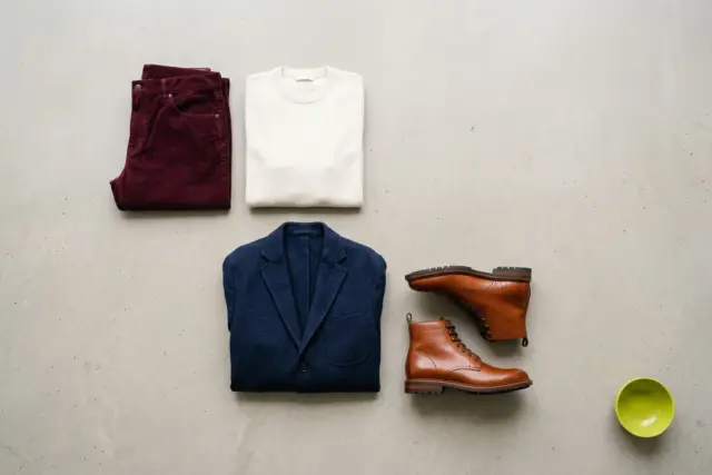 An overhead flat lay displays burgundy corduroy pants, an ivory merino crewneck, a navy wool blazer, and cognac leather boots neatly arranged on a light gray concrete floor, showcasing versatile fall