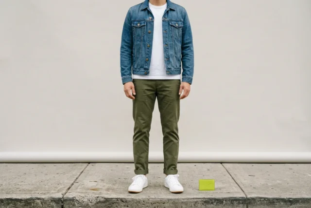 A person from the shoulders down, wearing a mid-wash denim jacket, open over a white cotton T-shirt, with olive cotton chinos and white leather sneakers, standing on a sidewalk.