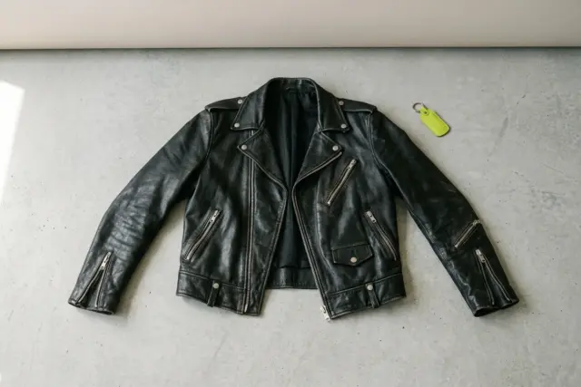 A black leather biker jacket, laid flat and open, its silver zippers and worn texture visible on a smooth, light grey concrete surface.