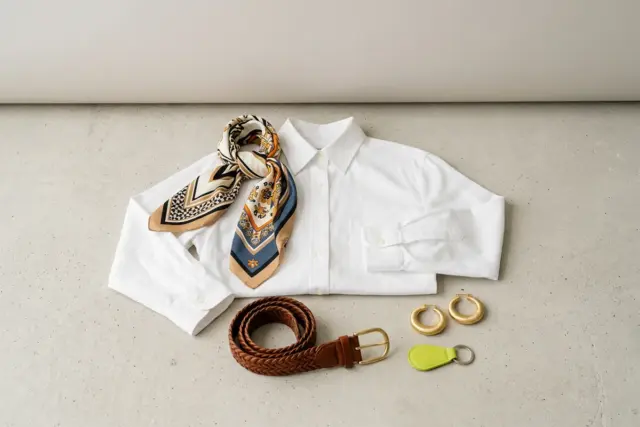 A crisp white cotton button-down shirt flat-laid on a light concrete surface, surrounded by a vibrant patterned silk scarf, a woven brown leather belt, and a pair of distinctive gold hoop earrings.