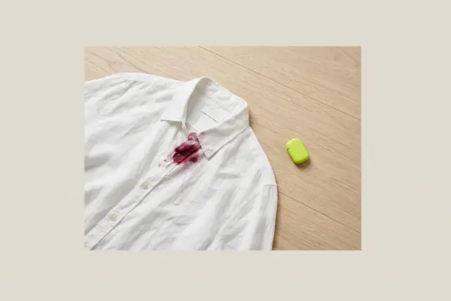 An overhead flat lay shows a crumpled white linen shirt with a fresh, vibrant crimson blood stain near the collar, resting on a clean light wood floor.