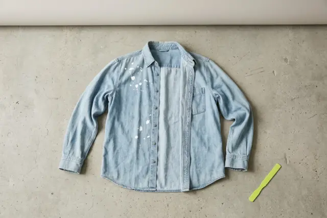 An unbuttoned pale blue denim shirt with a few fresh drops of white latex paint splattered near the collar, laid flat on a light grey concrete floor.
