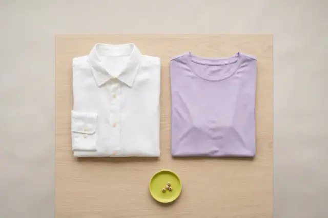 An overhead flat lay showcases a crisp white linen shirt and a soft lavender modal t-shirt, meticulously folded on a smooth, light beige wooden surface, awaiting gentle care.