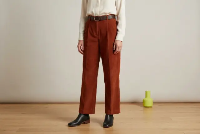From the shoulders down, a person wears rust wide-leg corduroy pants with an ivory silk-blend blouse tucked in and a dark brown leather belt, completed by black ankle boots on a smooth, light wooden f