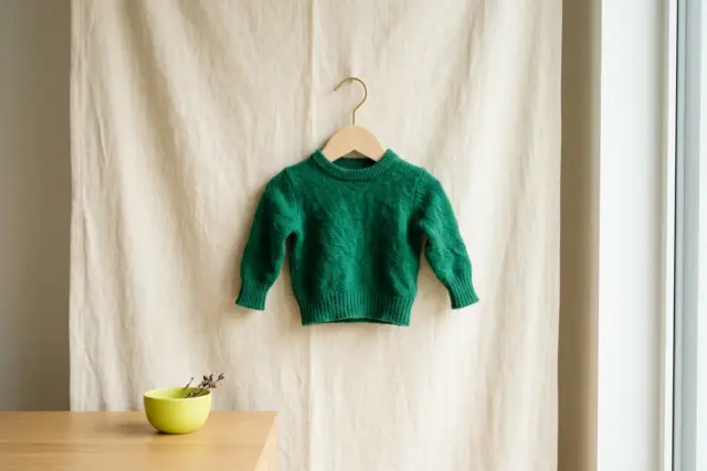 A tiny, noticeably shrunken emerald green merino wool sweater hangs on a slender wooden hanger against a soft cream fabric backdrop.