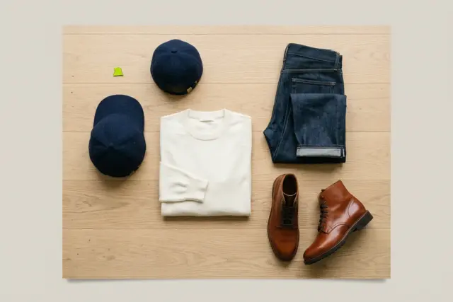 An overhead flat lay on a light wooden floor displays a neatly folded ivory merino crewneck with dark-wash denim jeans, a navy wool cap, and polished cognac leather boots.