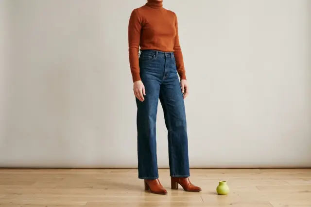 A person stands on a light wooden floor, wearing a fitted rust merino turtleneck tucked into dark-wash wide-leg jeans that neatly fall over cognac leather heeled boots with a pointed toe.
