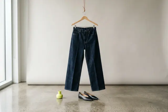 A pair of dark-wash wide-leg denim jeans hangs on a wooden hanger, with black leather pointed-toe flats sitting on a polished concrete floor beneath.