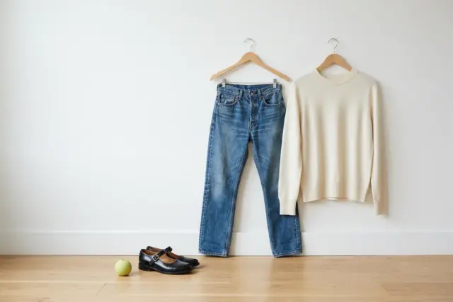 A black leather Mary Jane shoe sits on a light hardwood floor next to mid-wash straight-leg jeans and an ivory merino crewneck sweater, both neatly hung on simple wooden hangers against a clean white