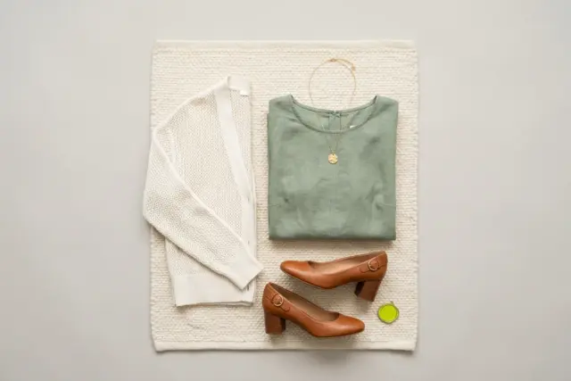 An overhead flat lay displays a neatly folded sage green midi dress, an ivory cotton-blend cardigan, cognac leather block heels, and a gold pendant necklace arranged on a textured cream rug.