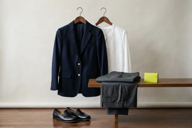 A navy blazer on a sleek wooden hanger, partially overlapping a white cotton-blend blouse on another hanger, with charcoal tailored trousers folded neatly on a shelf below, and black leather loafers r