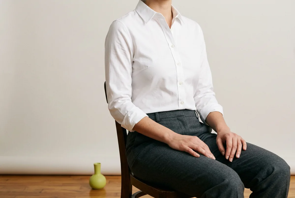 Person photographed from the shoulders down, wearing a classic white button-down shirt and grey trousers. They are seated, demonstrating that there is no gaping at the bust or pulling at the buttons.