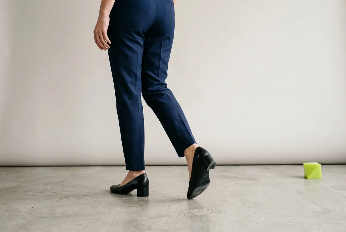 Person photographed from the waist down, walking away from the camera on a light gray concrete floor. They are wearing classic black leather pumps and navy blue tailored trousers. The focus is on the