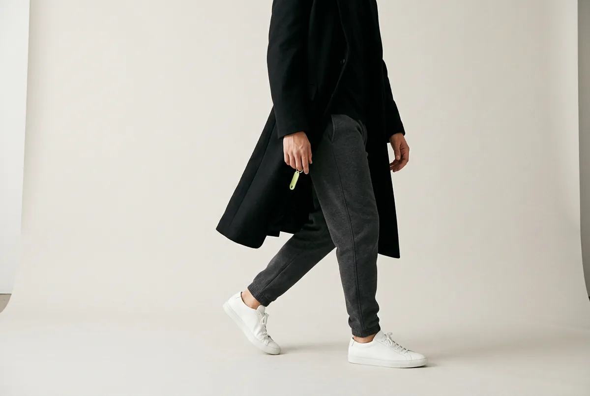 Person photographed from the waist down, walking. They are wearing dark gray tapered sweatpants, clean white sneakers, and a long black wool coat that is open.