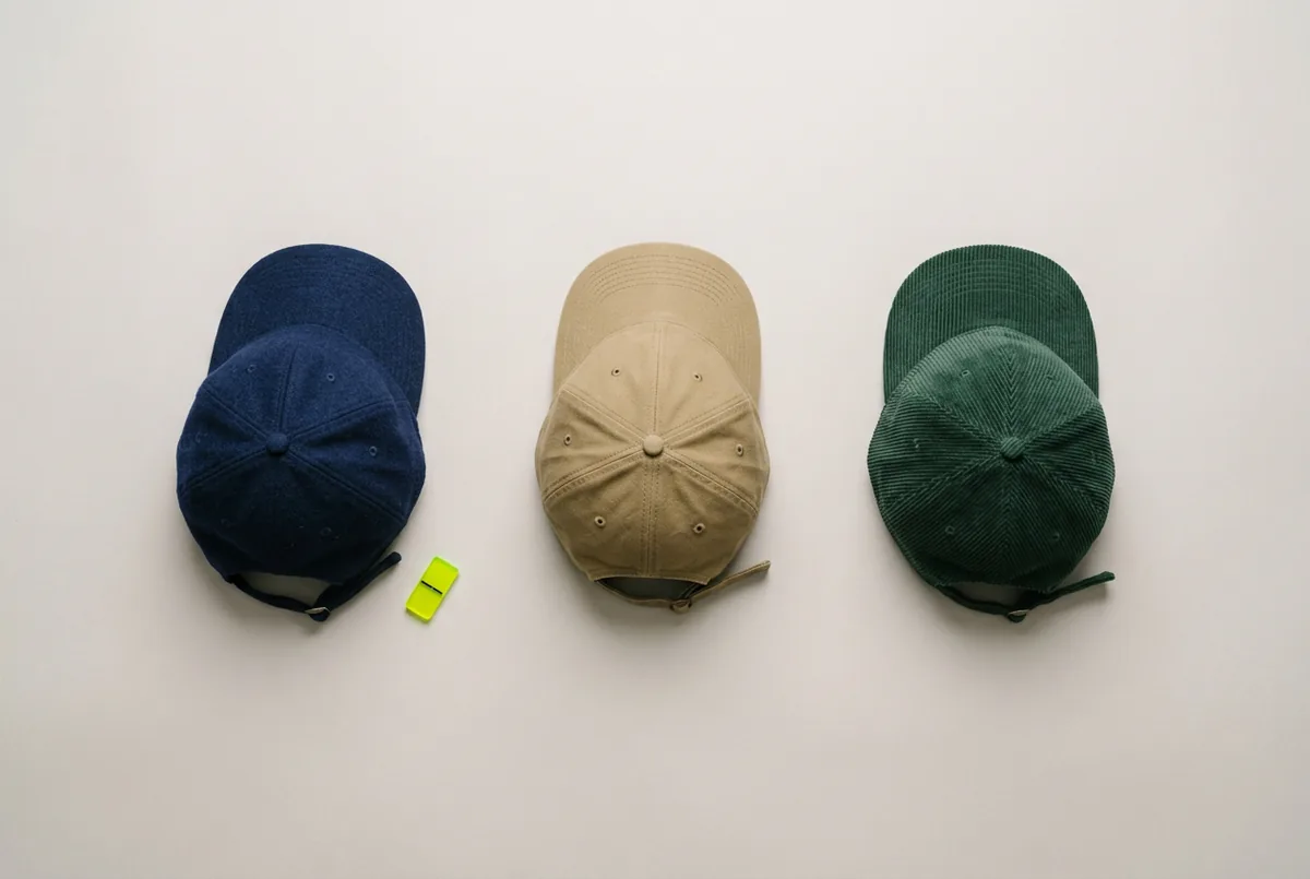 Overhead flat lay of three baseball caps on a neutral background: one in navy wool, one in khaki canvas, and one in dark green corduroy.