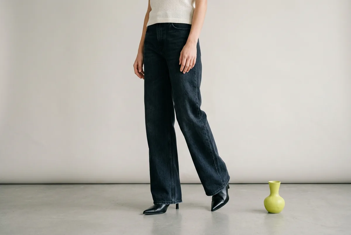 Person photographed from the waist down, wearing dark-wash full-length wide-leg jeans and pointed-toe heeled boots in black leather. Only the toe and front of the boot is visible.
