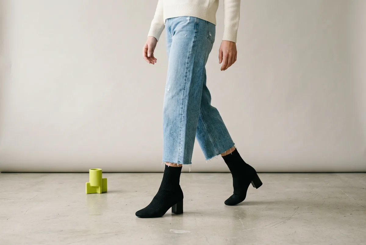 Person photographed from the knees down, wearing cropped light-wash wide-leg jeans and black sock boots that fit snugly at the ankle.