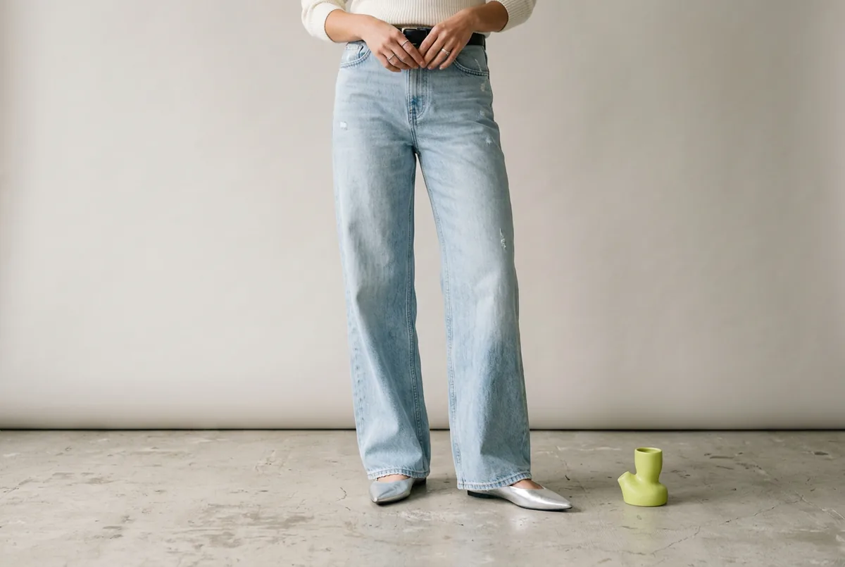Person photographed from the waist down wearing full-length, light-wash wide-leg jeans and silver pointed-toe flats, standing on a pale gray concrete floor.